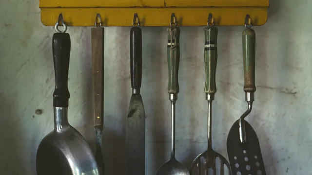 A small pan, meat fork, icing spatula, spoons and a slotted spatula hanging from hooks under a yellow wooden shelf