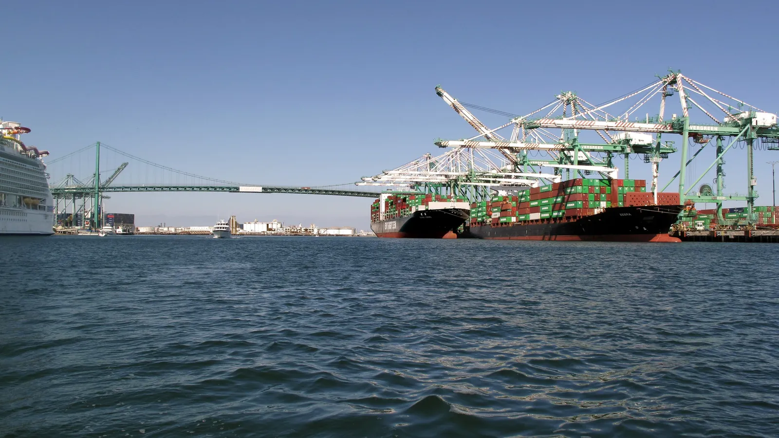 Stacked shipping containers on cargo ships docked at the Port of Los Angeles beneath a green suspension bridge