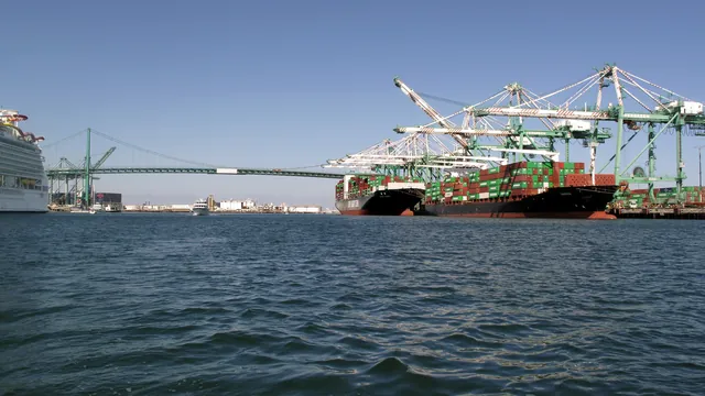 Stacked shipping containers on cargo ships docked at the Port of Los Angeles beneath a green suspension bridge