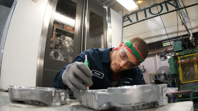 A machinist in safety glasses inspects an aluminum die-casting next to a CNC machine at Port City Castings in Muskegon, Michigan