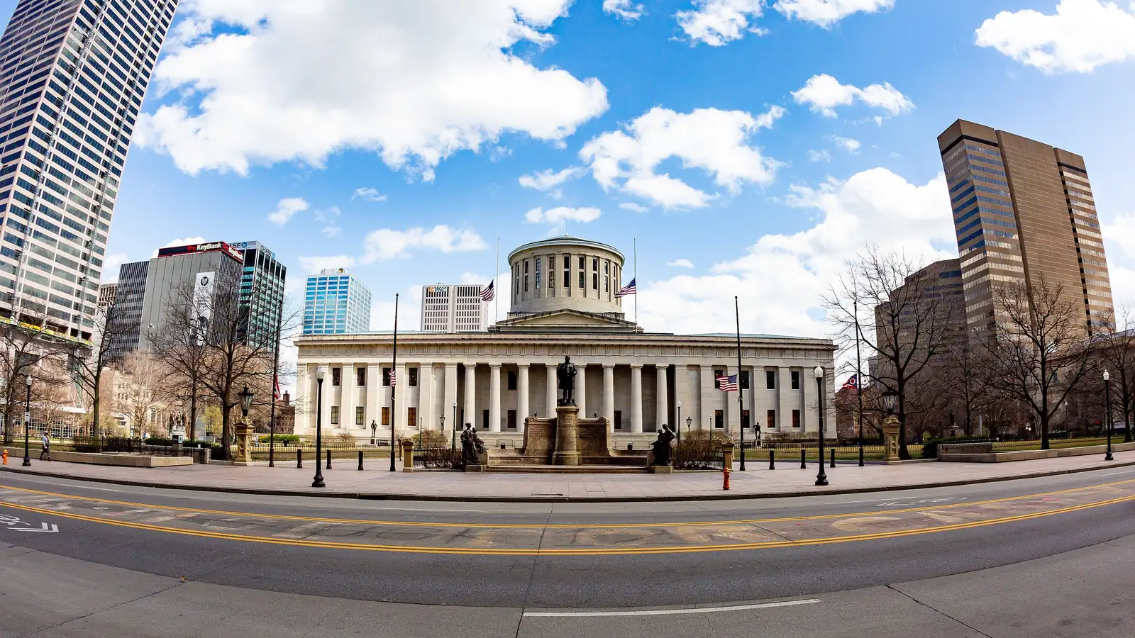 The columned Ohio Statehouse and its plaza under a blue sky, framed by downtown Columbus office towers