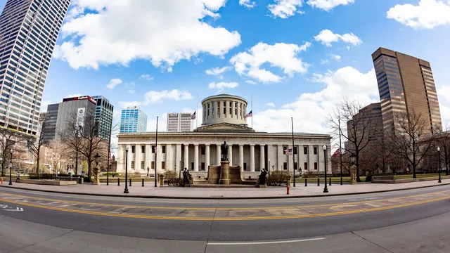 The columned Ohio Statehouse and its plaza under a blue sky, framed by downtown Columbus office towers