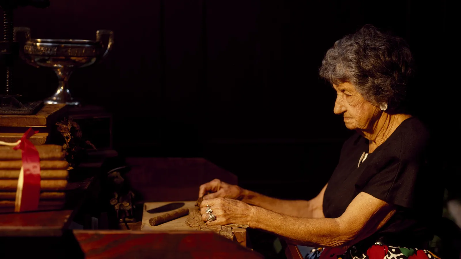 An older woman hand-rolling cigars at a wooden workbench in a dimly lit factory in Tampa, Florida