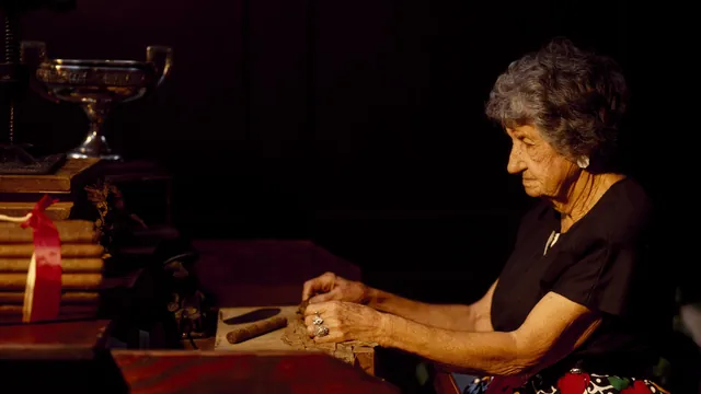 An older woman hand-rolling cigars at a wooden workbench in a dimly lit factory in Tampa, Florida